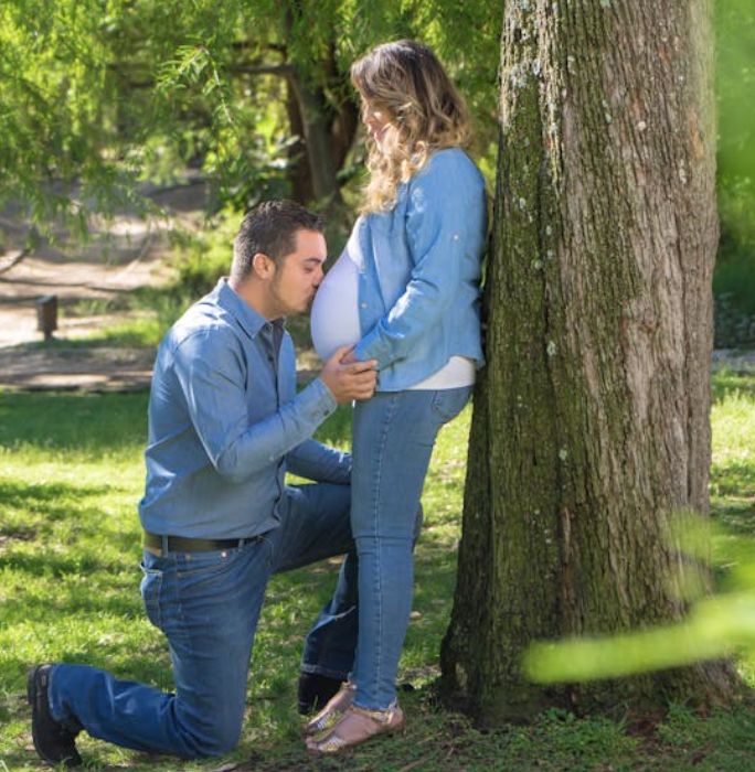 husband kissing wife's pregnant belly while kneeling on one kneww in the park against a tree